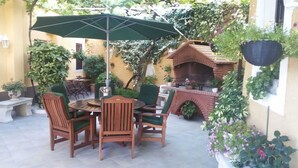 A patio with a round wooden table, four chairs, a green umbrella, and a brick barbecue grill.