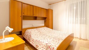 A bedroom with a bed, wooden wardrobe, bedside table, and a window with sheer curtains.