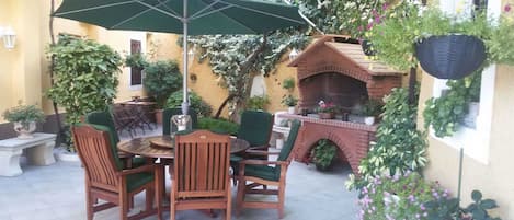 Eine Terrasse im Freien mit einem runden Holztisch und Stühlen, einem grünen Sonnenschirm und einem Grill aus Ziegelsteinen.