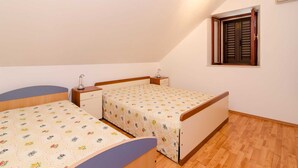 A bedroom with two single beds, a small bedside table with a lamp, a window with shutters, and an air conditioning unit on the wall.