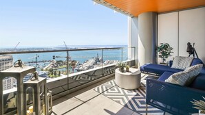 A balcony with a view of a marina and a bridge, furnished with a blue sofa, a round coffee table, and potted plants.