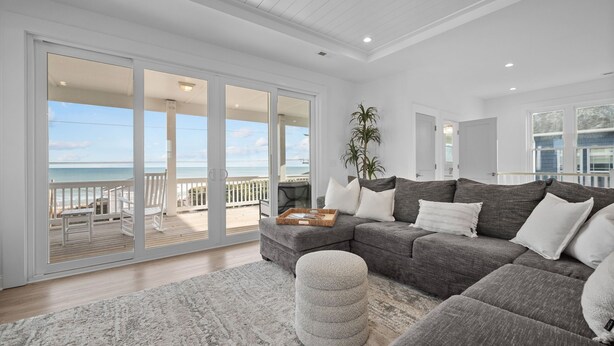 A spacious living room with a large gray sectional sofa, a round ottoman, and a view of the ocean through sliding glass doors. - Luxe 4 Bedroom Home w/ Ocean views & Quick Private Beach Access in Surf City! (Surf City)