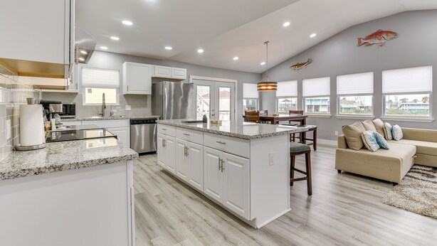 A modern kitchen with white cabinets, a granite countertop, and stainless steel appliances. There is a dining area with a wooden table and chairs, and a beige sofa in the living room. - Modern Freeport Home w/ Views: Walk to Beach! (Freeport)