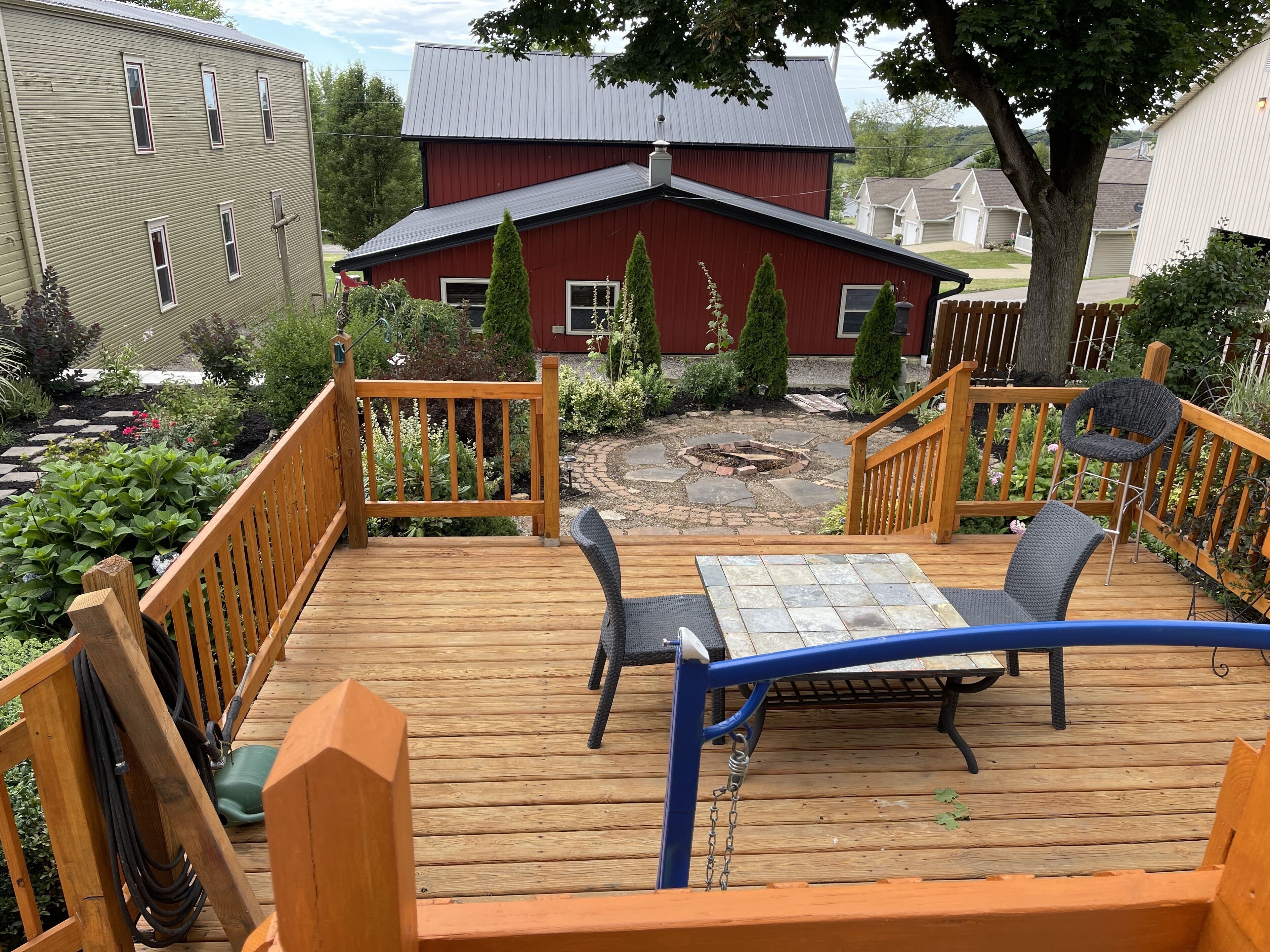 Une terrasse en bois avec une table et des chaises, entourée d’un jardin et, au fond, d’une maison rouge.