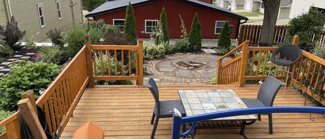 Une terrasse en bois avec une table et des chaises, entourée d’un jardin et, au fond, d’une maison rouge.
