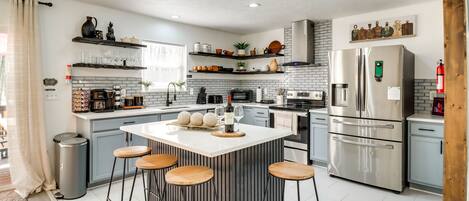 A modern kitchen with a central island, bar stools, stainless steel appliances, and decorative items on shelves.