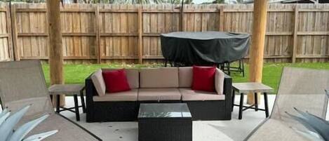 An outdoor patio with a thatched roof, a sofa, two chairs, and a small table.