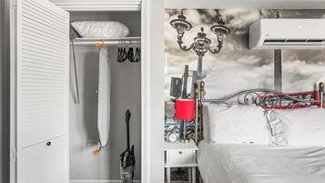 A bedroom with a bed, a nightstand, a red lamp, an air conditioner, and a closet with hanging clothes.