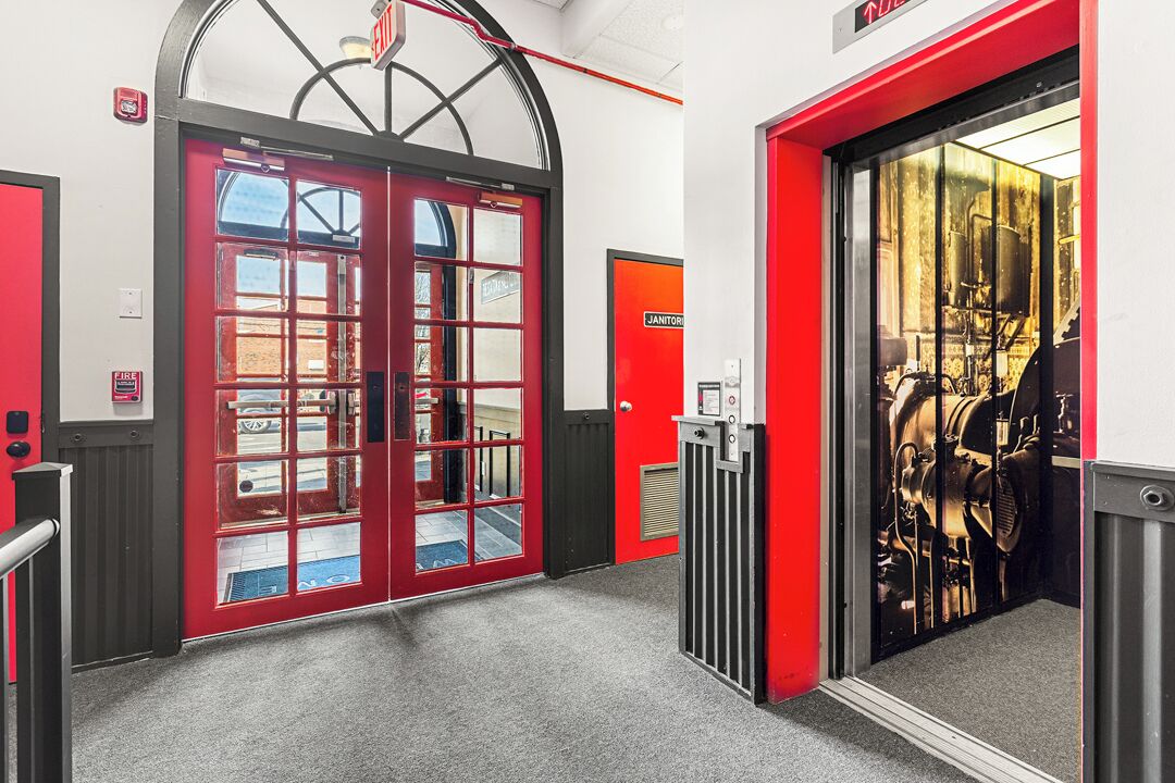 A fire station entrance with red doors, a fire alarm, and a view of fire equipment through a glass door.