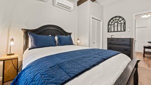 A bedroom with a bed, a dresser, a nightstand, an air conditioning unit, and a door.