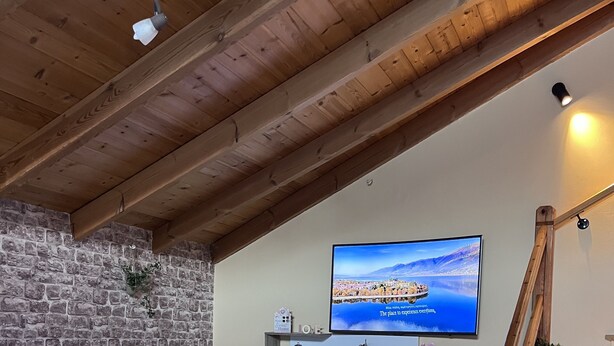 Un salon chaleureux avec un mur en pierre, un plafond en bois, un téléviseur fixé au mur, un canapé, une petite table avec des bougies et un escalier.