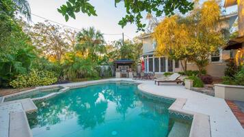 A swimming pool with a patio area, surrounded by lush greenery and a house in the background.