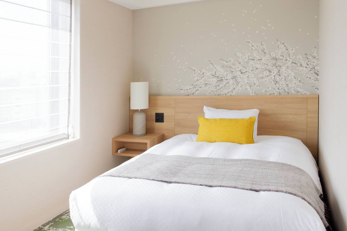 Un lit bien fait, avec des oreillers blancs et jaunes, une lampe de chevet et un mur orné d’une fresque florale.