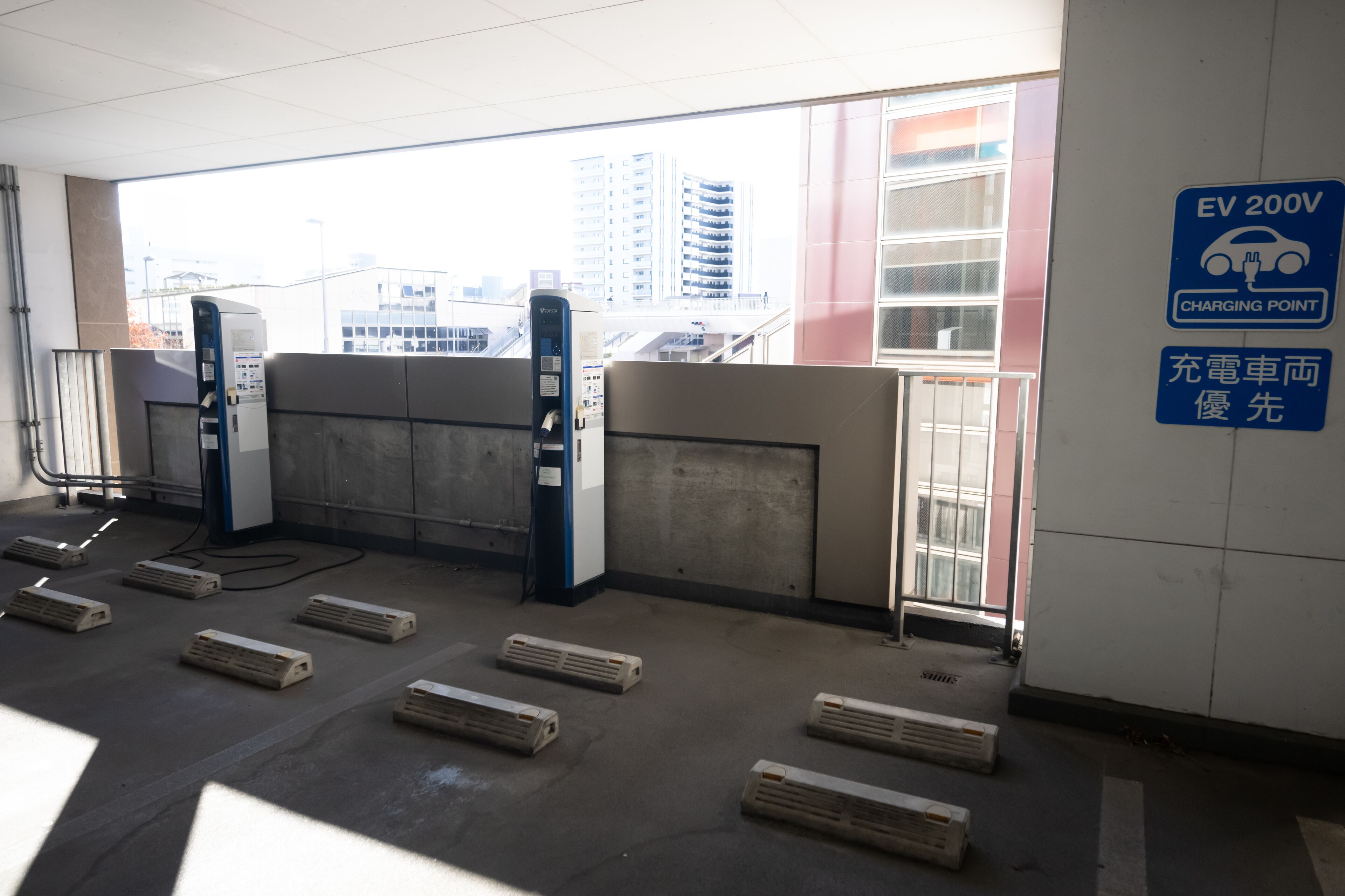 Señal de punto de carga para vehículos eléctricos de 200V, barreras de hormigón y vistas de edificios a través de una ventana.