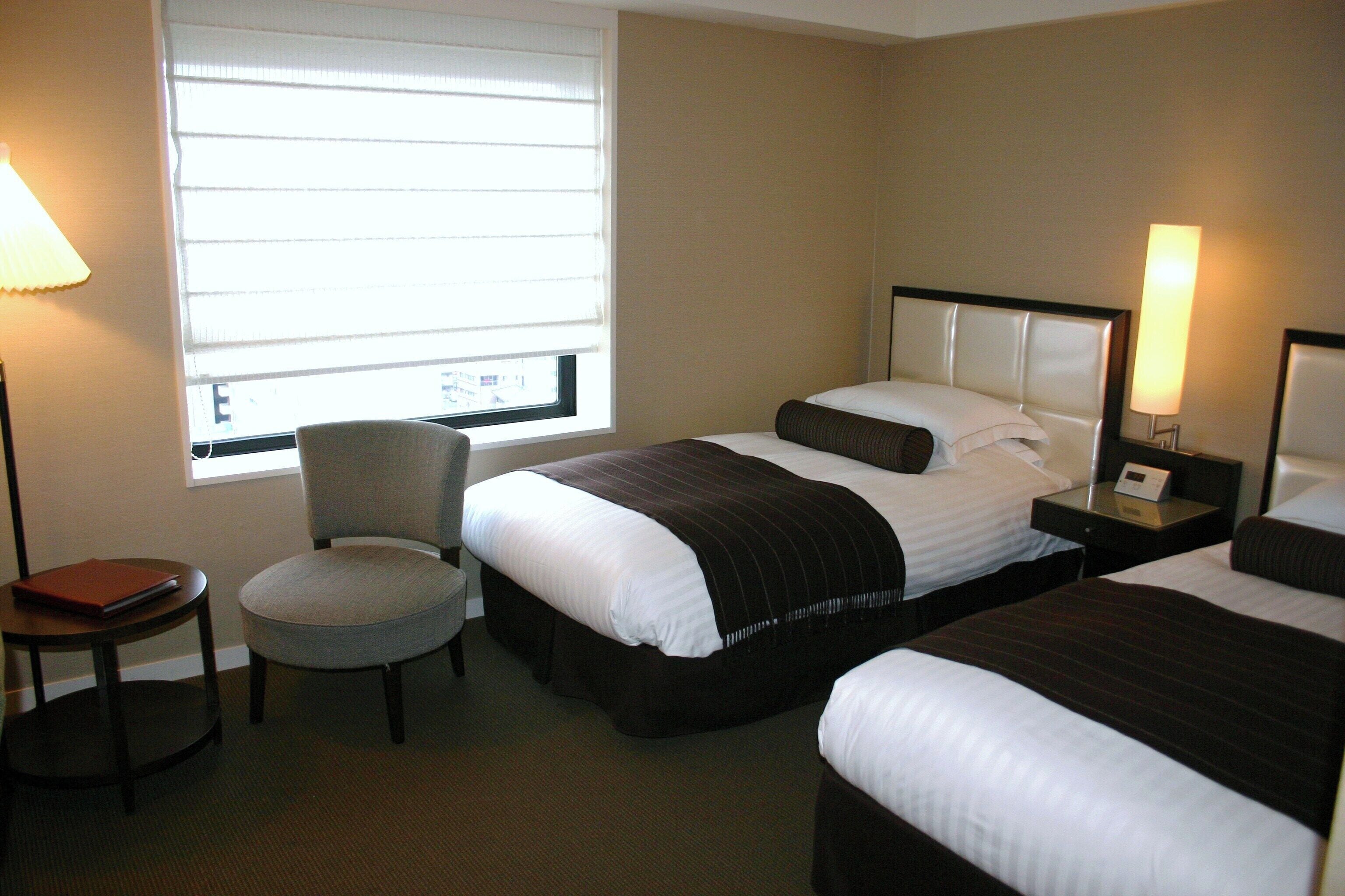 Une chambre d’hôtel avec deux lits, une chaise, une petite table et une fenêtre avec des stores.