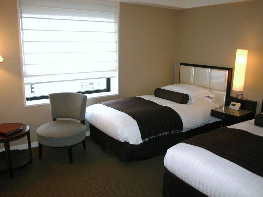 Une chambre d’hôtel avec deux lits, une chaise, une petite table et une fenêtre avec des stores.