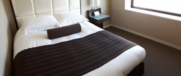 Una cama bien hecha con una colcha a rayas, almohadas blancas y una almohada cilíndrica marrón oscuro. Hay una mesita de noche con una lámpara y un cartel.