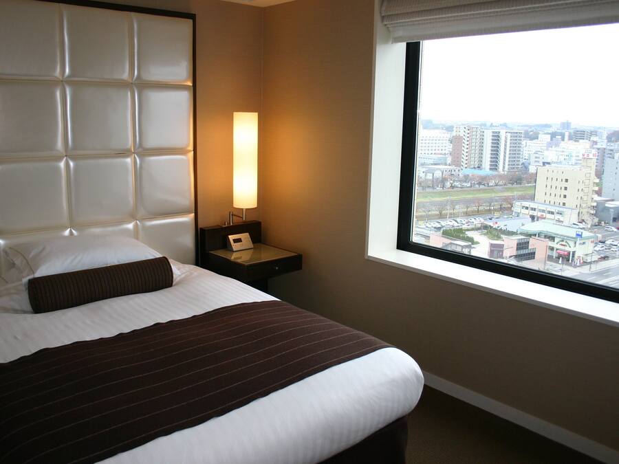 Ein Hotelzimmer mit einem großen Bett, einem Nachttisch mit Lampe und einem Fenster mit Blick auf die Stadt.
