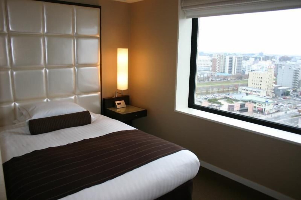 A hotel room with a large bed, a nightstand with a lamp, and a window with a city view.