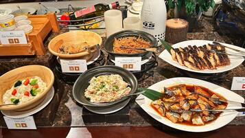 A buffet table with various dishes including a bowl of potato salad, a bowl of vermicelli salad, a bowl of rice with vegetables, a plate of grilled fish, and a bottle of cold wine.