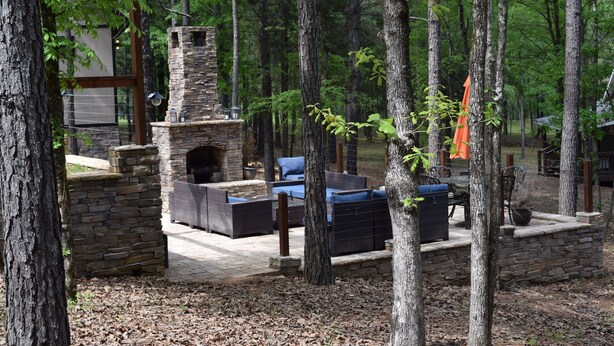 Une cheminée en pierre, entourée d’un espace de détente extérieur, située dans un endroit boisé, agrémenté d’arbres et d’un mur en pierre.