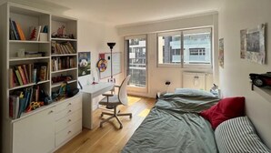 A bedroom with a bed, a desk with a chair, a bookshelf, and a window with a view of buildings.