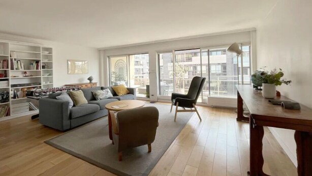 A spacious living room with a grey sofa, a round coffee table, and a wooden dining table. There is a bookshelf filled with books and decorative items. Large windows provide natural light and a view of the outside.