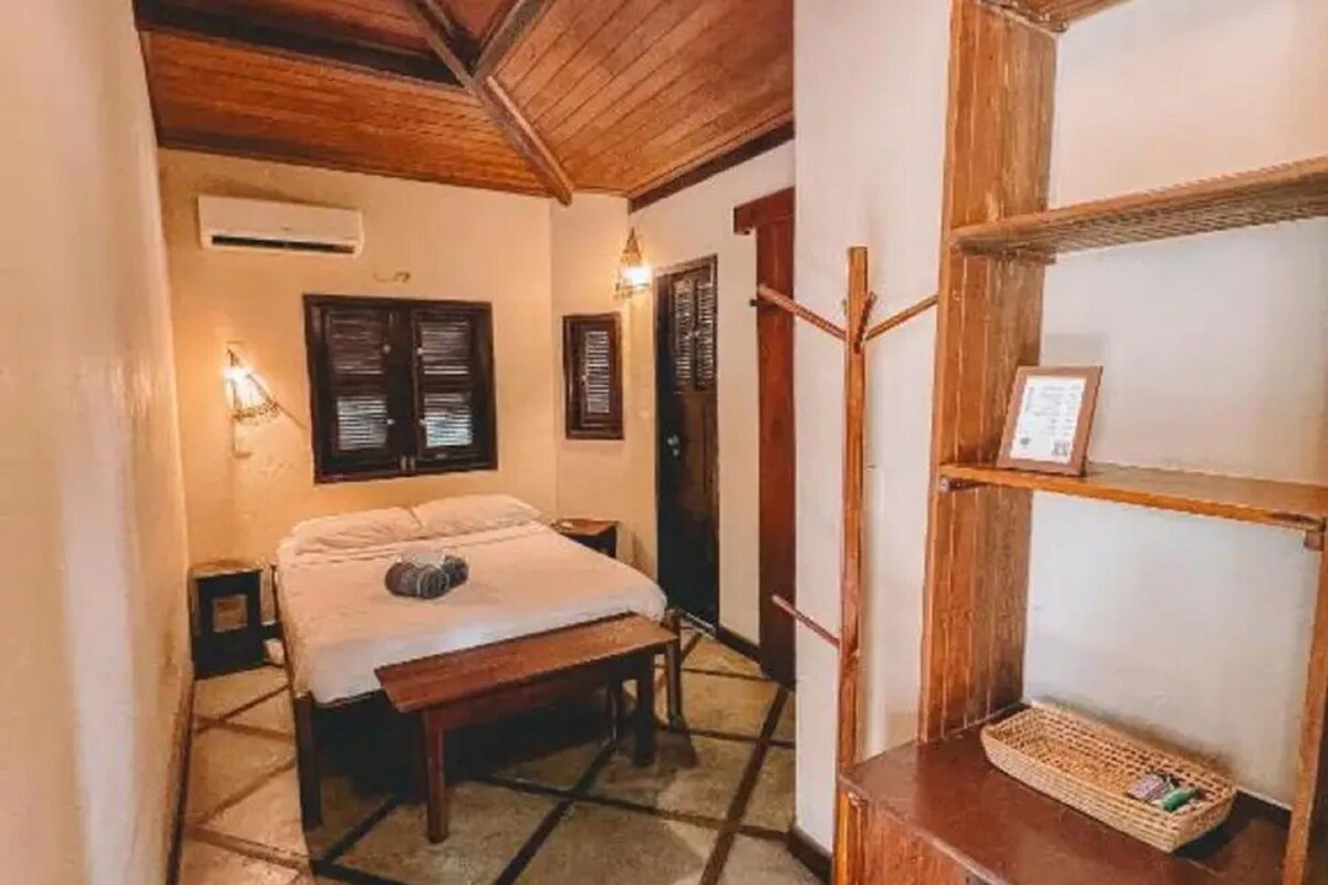 Une pièce comprenant un lit, un banc en bois, une étagère fixée au mur et une porte.