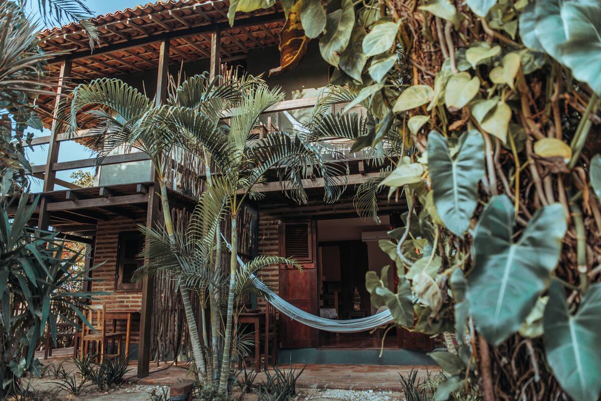 Una casa de madera con balcón, rodeada de plantas tropicales y una hamaca.