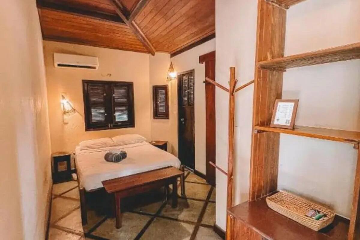 Una habitación con una cama, un banco de madera, un estante en la pared y una puerta.