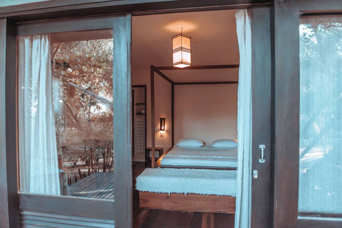 Una habitación con cama, piso de madera, lámpara colgante y vista a árboles a través de un ventanal.