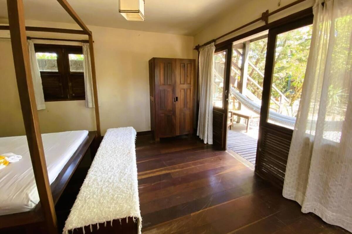 Una habitación con una cama de madera, un banco, un armario y una ventana con cortinas.