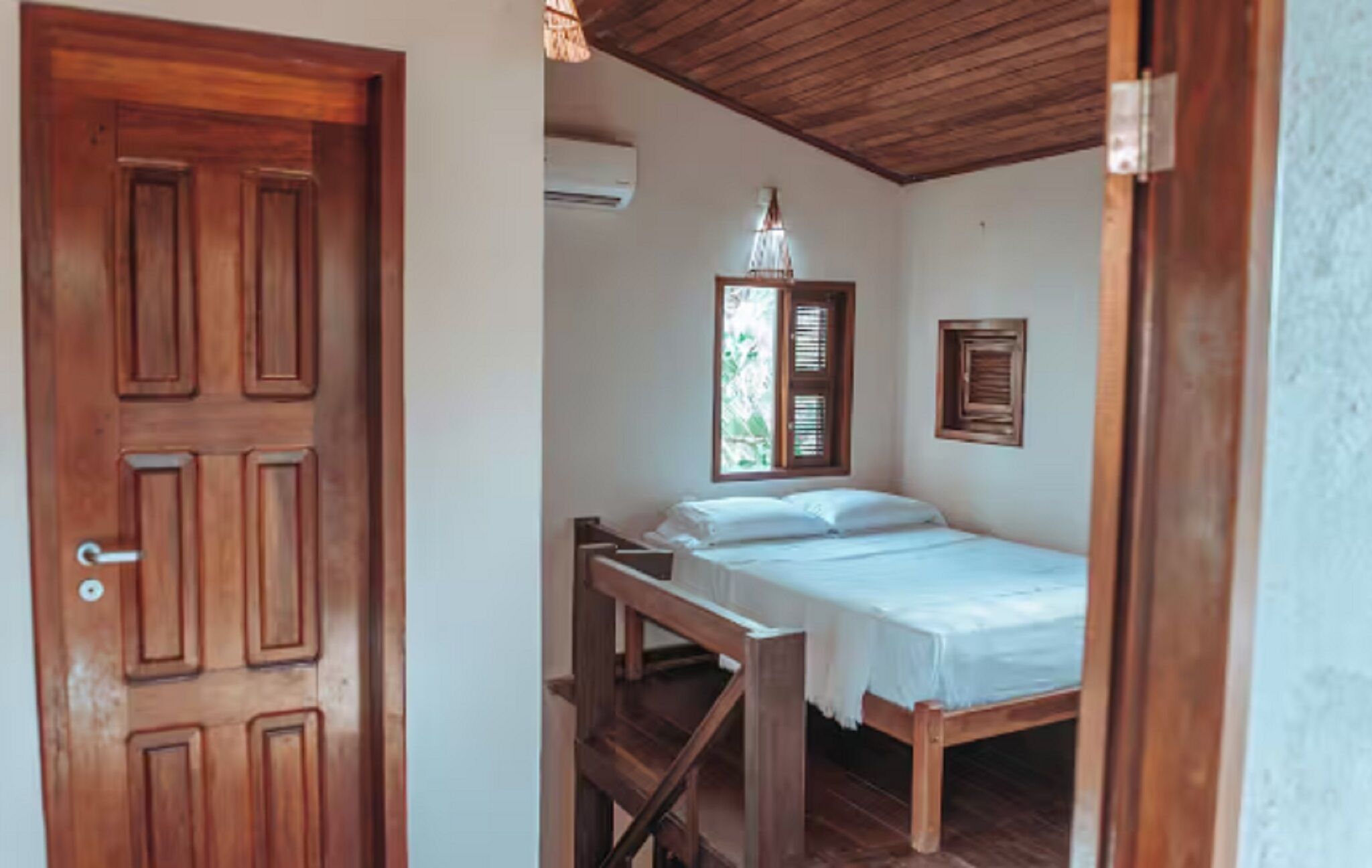 Una habitación con puerta de madera, una cama con ropa blanca, un banco de madera, una lámpara colgante y una ventana con contraventanas.
