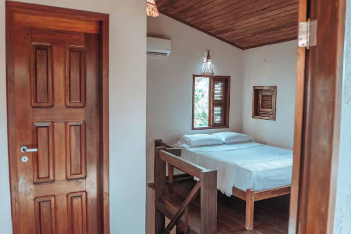 Una habitación con puerta de madera, una cama con ropa blanca, un banco de madera, una lámpara colgante y una ventana con contraventanas.