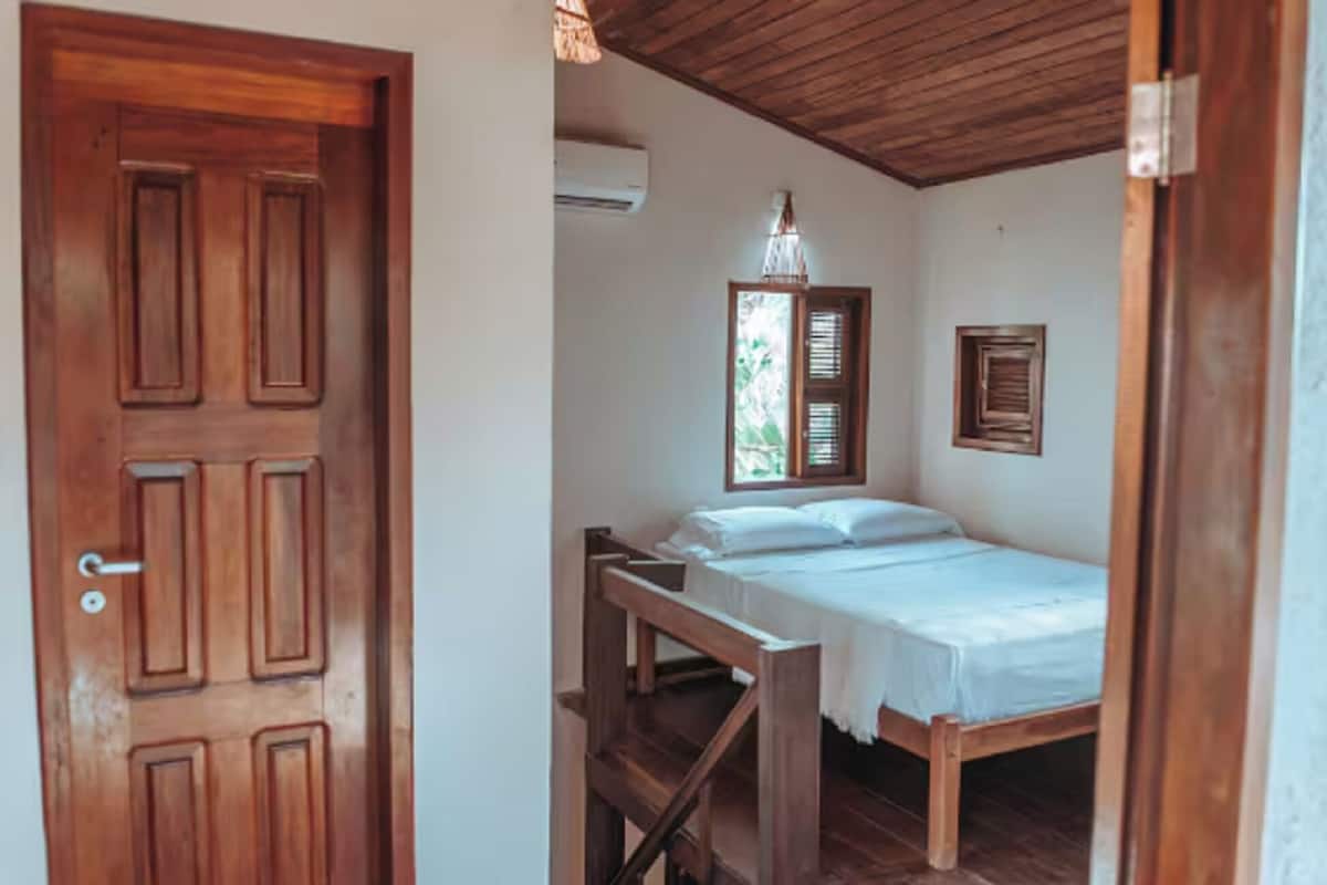 A room with a wooden door, a bed with white bedding, a wooden bench, a hanging light fixture, and a window with shutters.