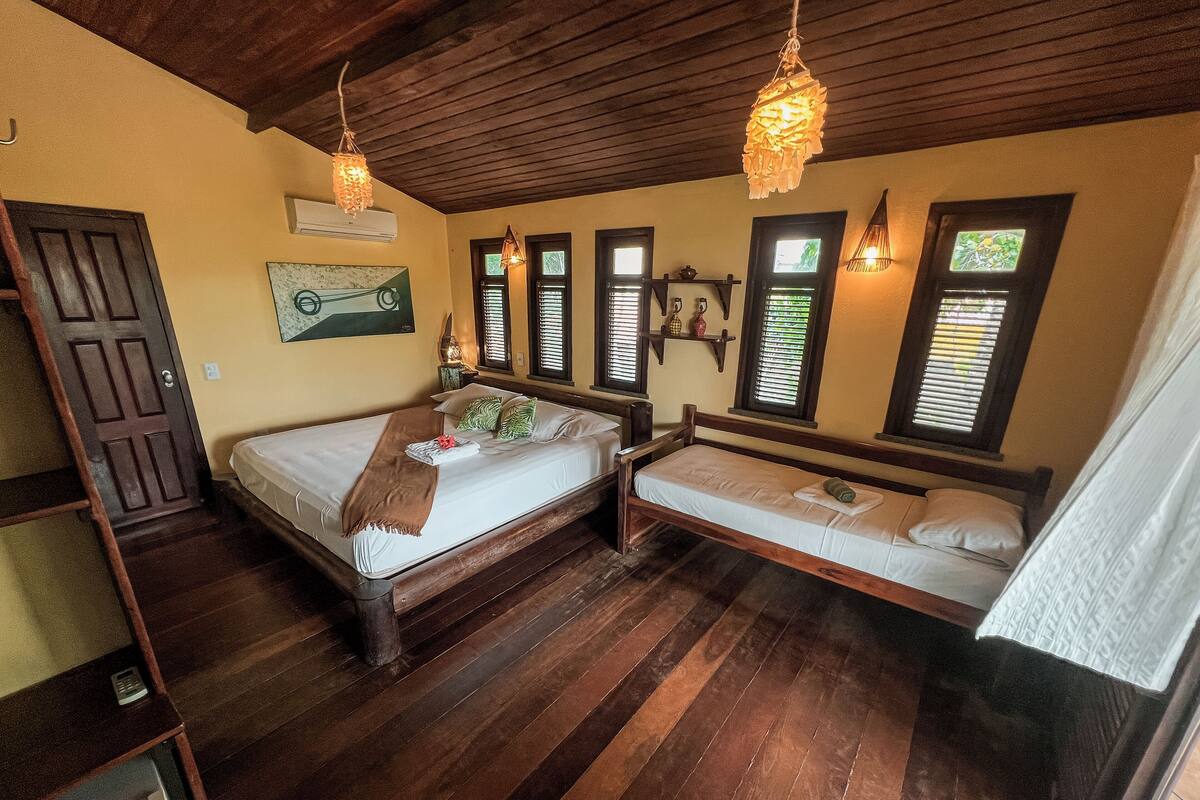 Habitación con una cama de madera, una cama individual, un escritorio y una silla. Hay ventanas con persianas, una lámpara colgante y un estante en la pared con adornos.