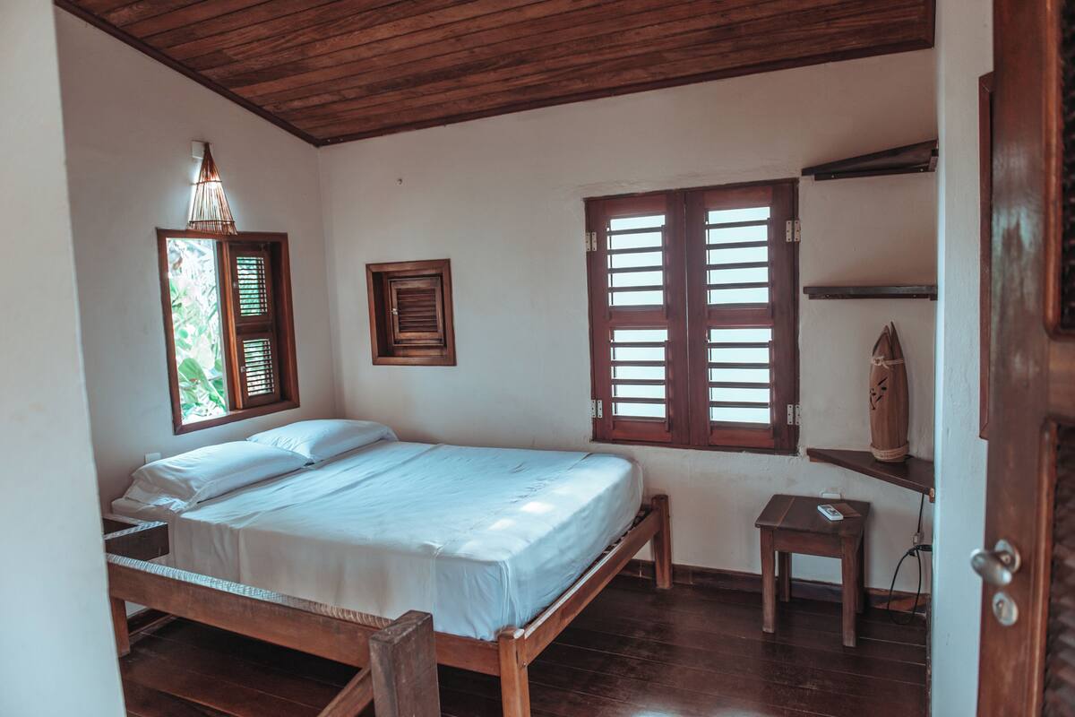 Un dormitorio con cama de madera, una mesita de noche pequeña, una mesita auxiliar, un estante de madera y una ventana con contraventanas.