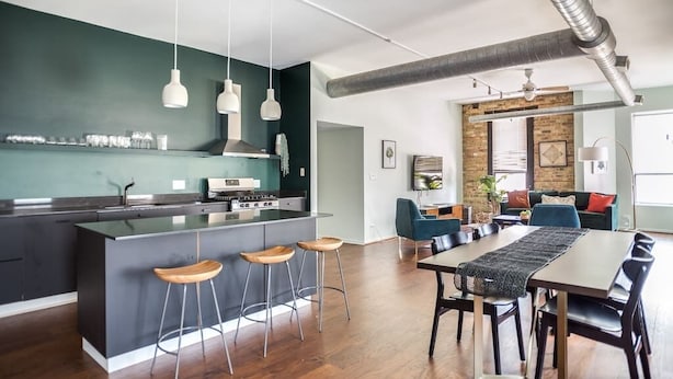 A modern kitchen with a central island, bar stools, pendant lights, and a dining area with a long table and chairs.