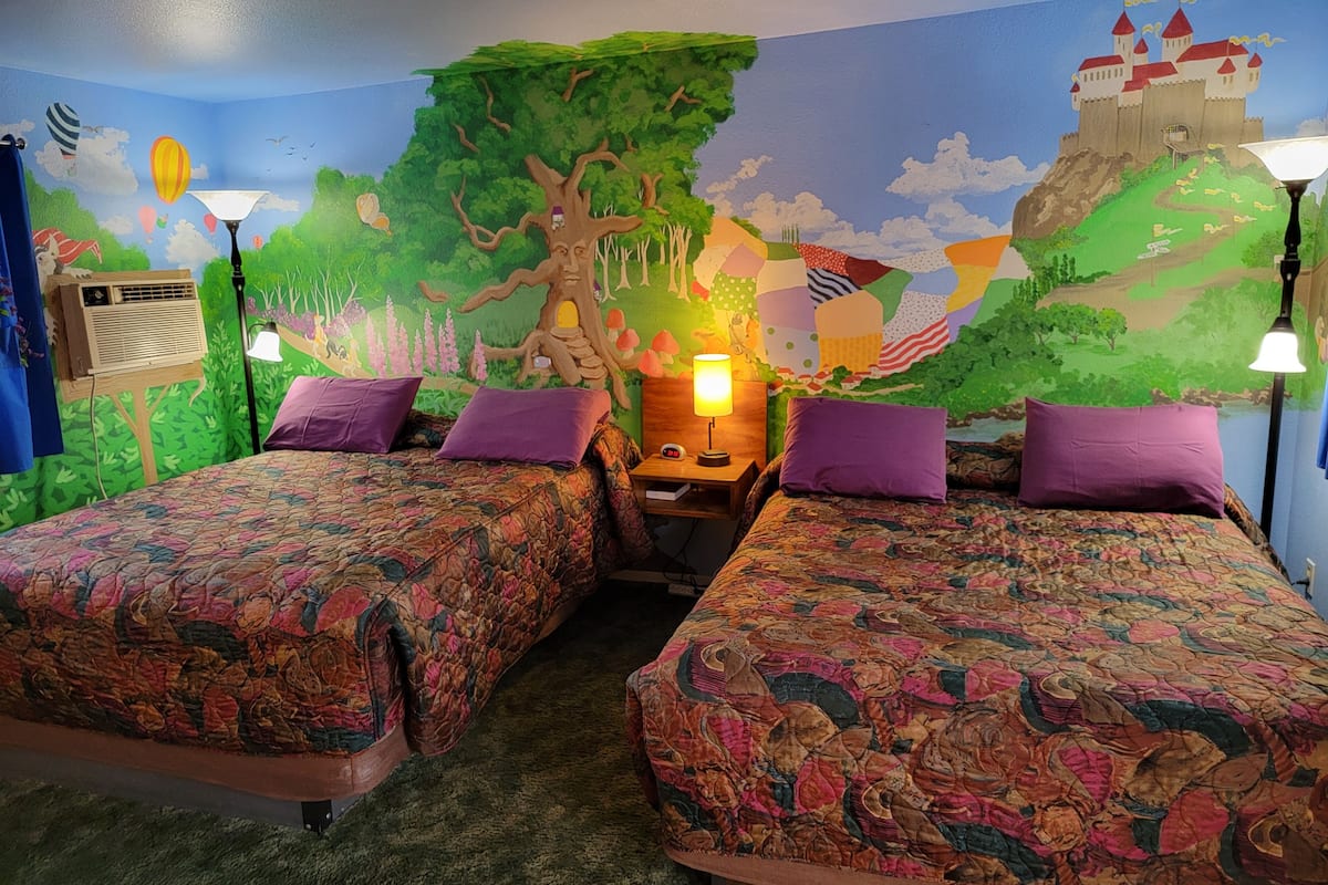 A room with two beds, a nightstand with a lamp, and a mural of a castle and tree.