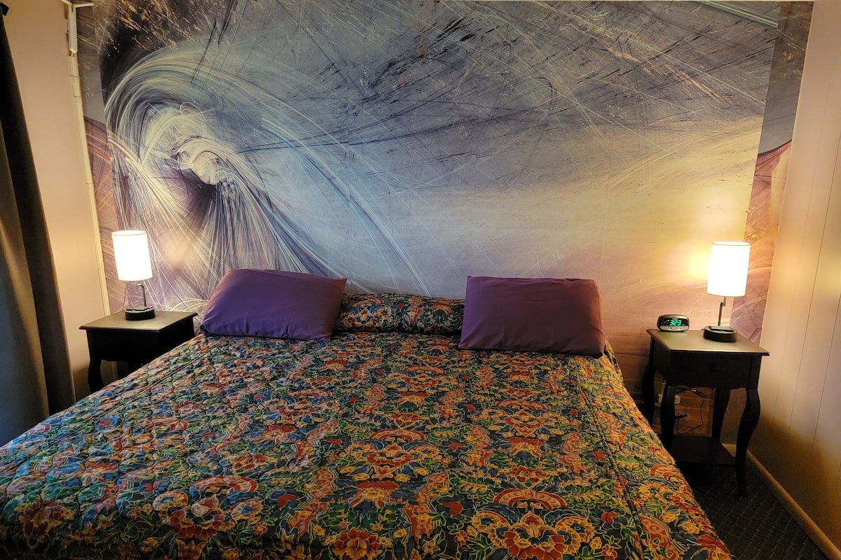 A hotel room with a large bed, two nightstands, two lamps, and a colorful tapestry on the wall.