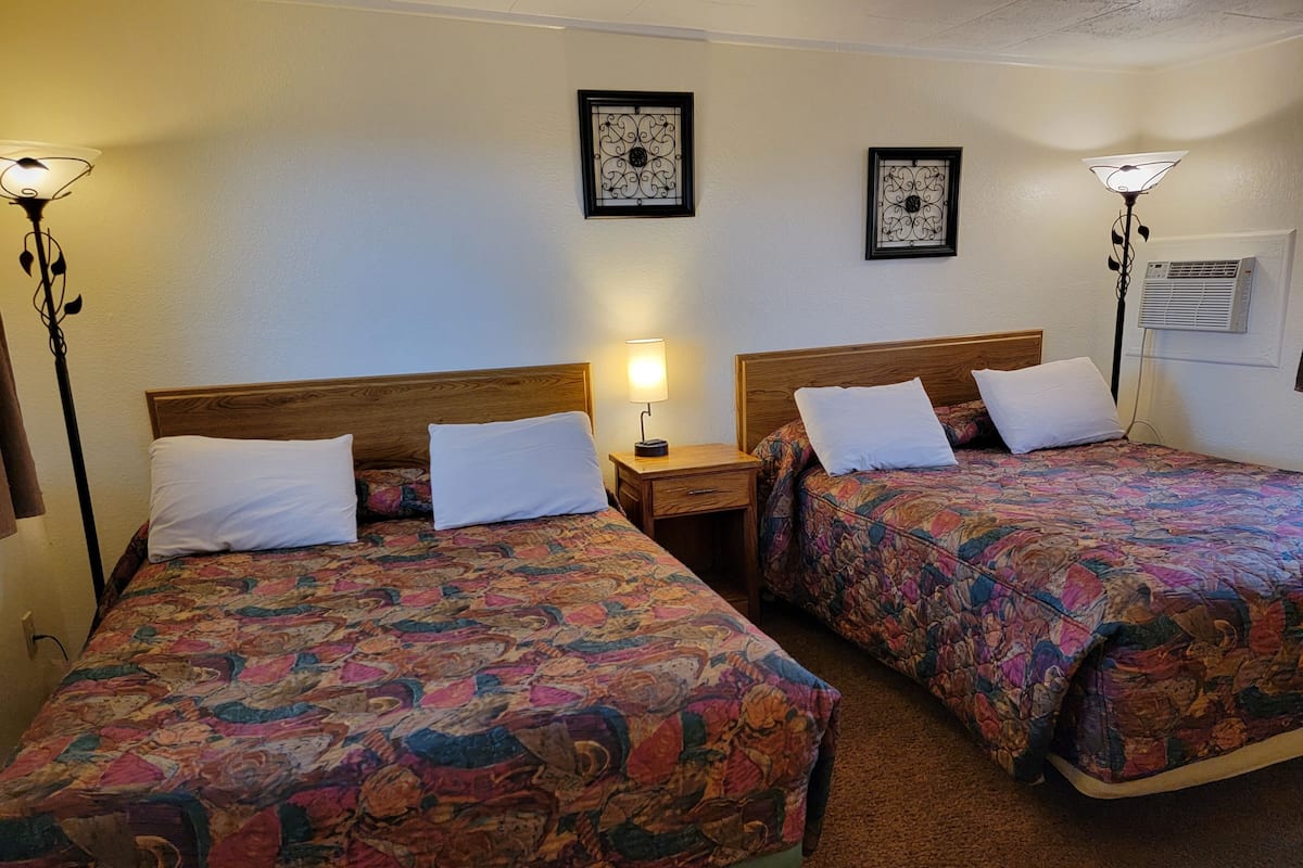 A hotel room with two beds, each with a floral bedspread and white pillows. There are two wall-mounted picture frames, a nightstand with a lamp, and a window with an air conditioning unit.