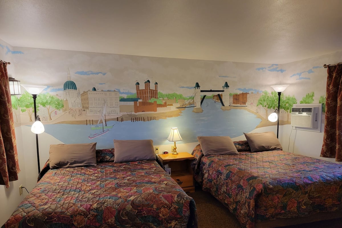 A room with two beds, a mural of a cityscape, a lamp, and an air conditioning unit.