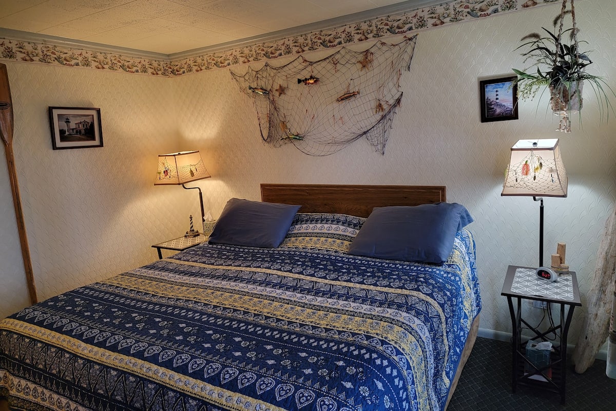 A bedroom with a patterned bedspread, a wooden headboard, a nightstand with a lamp, and a wall-mounted picture.