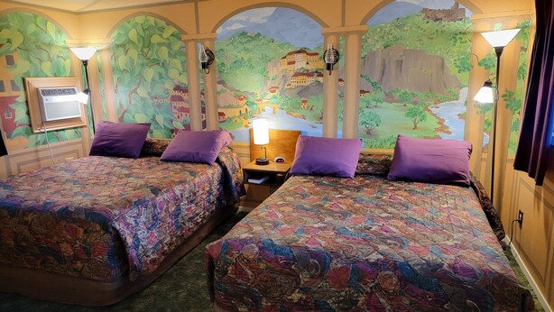 A hotel room with two beds, each with a patterned bedspread and purple pillows. There is a nightstand with a lamp and a wall-mounted air conditioner. The room features a mural of a landscape with trees and buildings. - La Collage Inn (Odessa)