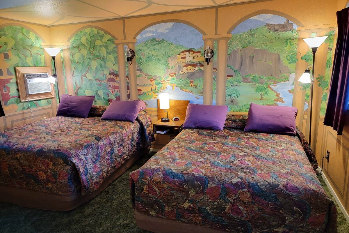 A hotel room with two beds, each with a patterned bedspread and purple pillows. There is a nightstand with a lamp and a wall-mounted air conditioner. The room features a mural of a landscape with trees and buildings.