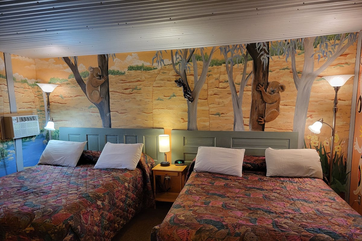 A room with two beds, each with a floral-patterned bedspread and white pillows. There is a nightstand with a lamp between the beds. The walls feature a mural of trees and a desert landscape.