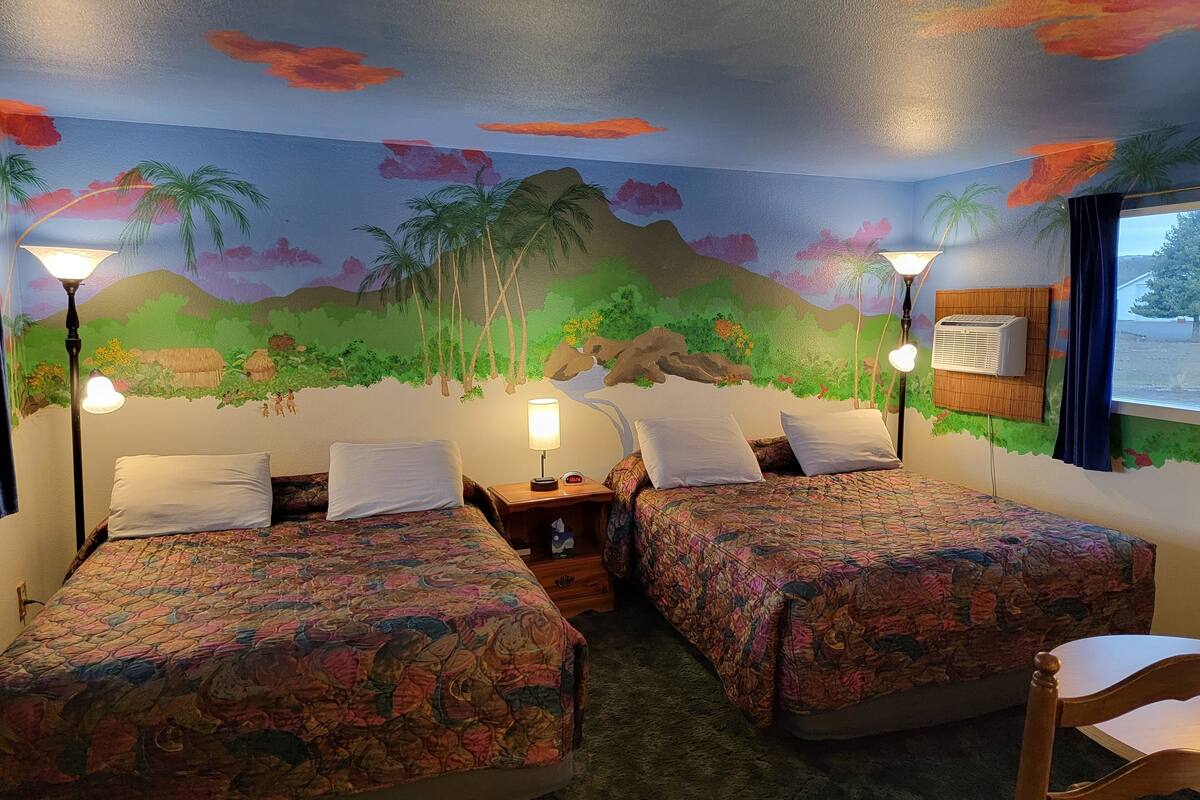 A room with two beds, each with a floral bedspread, a nightstand, a lamp, and a wall mural depicting a tropical scene.