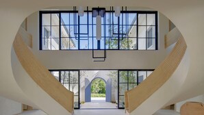 A spacious interior with a central staircase leading to an upper level, large windows, and a view of greenery outside.