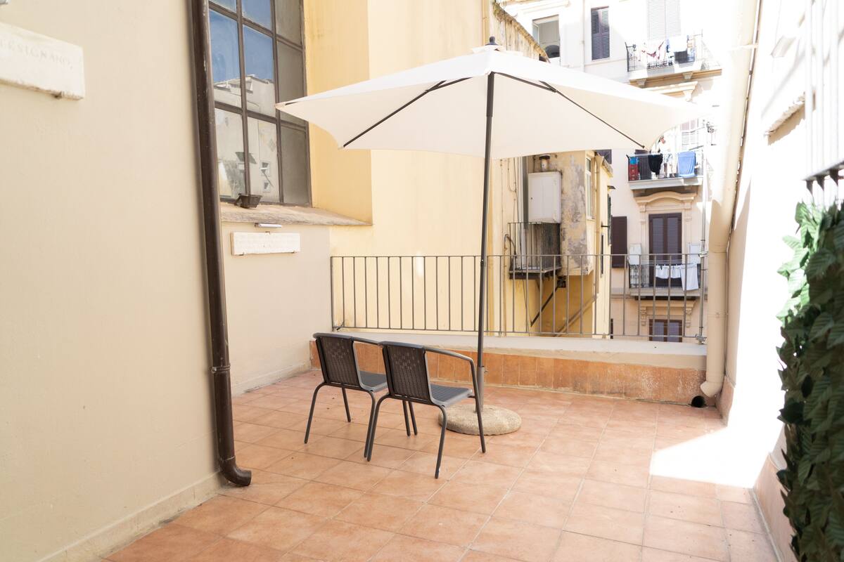 A small outdoor seating area with two chairs and a white umbrella, a tiled floor, and a view of a narrow street with buildings.