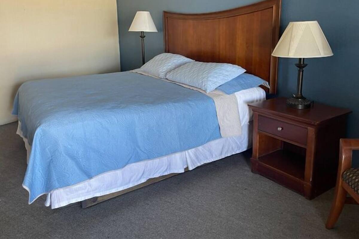 A neatly made bed with a blue bedspread and white pillows, flanked by two bedside tables with lamps, in a room with blue walls and a wooden headboard.